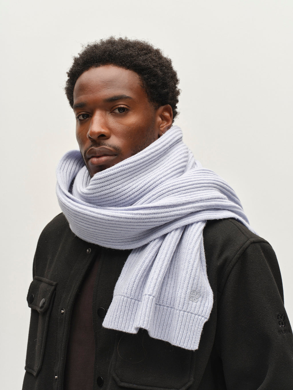 A man with short curly hair wears the Gabba GABeast Flash Scarf in Dusty Blue, wrapped around his neck, along with a black jacket, standing against a plain light background.