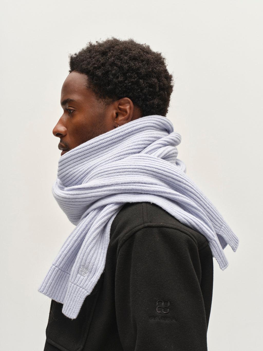 A person in a black coat is shown in profile wearing the Gabba GABeast Flash Scarf in Dusty Blue, a thick rib-knit accessory wrapped around their neck. The background is plain and light-colored.