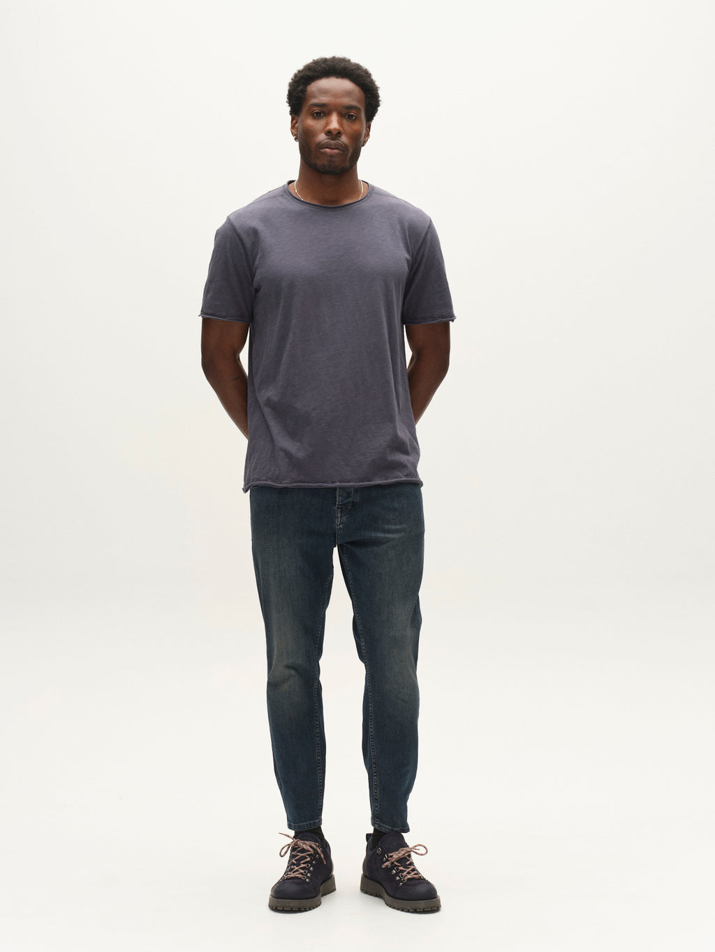 A man stands facing forward with his hands behind his back, wearing a dark gray t-shirt, dark sneakers, and Gabba’s GABalex Cropped Nham RS2113 Jeans in dark blue denim—crafted in a relaxed tapered fit from performance comfort ISKO fabric—against a plain light background.