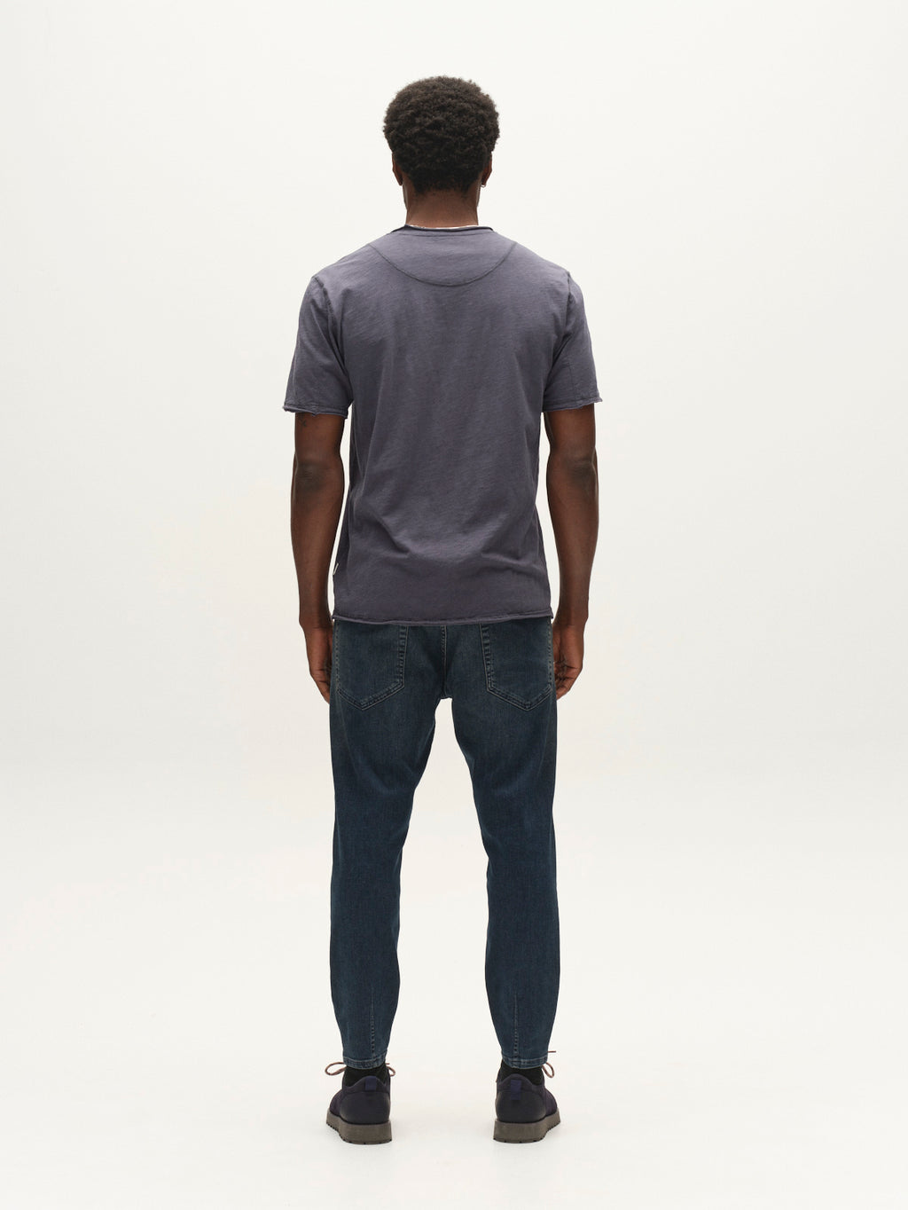 A man with short curly hair, wearing a dark short-sleeve shirt, Gabba GABalex Cropped Nham RS2113 Jeans in dark blue denim featuring a relaxed tapered fit and crafted from performance comfort ISKO fabric, and dark shoes stands facing away on a plain white background.