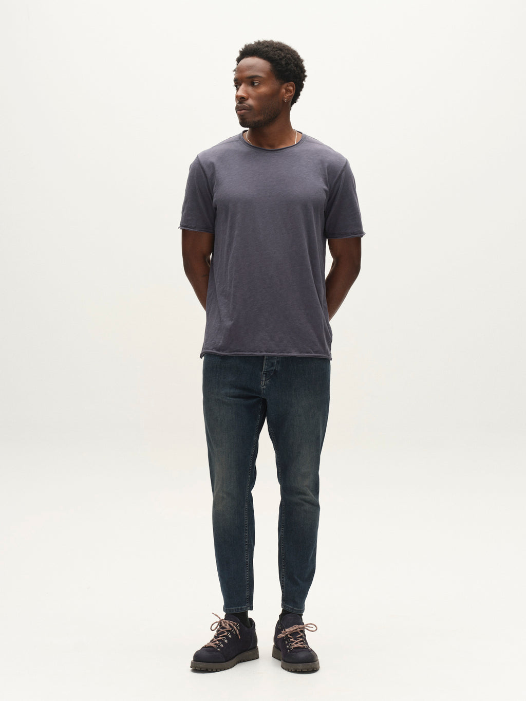 A man in a plain dark T-shirt, Gabba GABalex Cropped Nham RS2113 Jeans in dark blue denim, and dark shoes stands against a white background, facing slightly to the right with his hands behind his back. His jeans, expertly crafted from performance comfort ISKO denim by Gabba, offer both style and ease for any occasion.