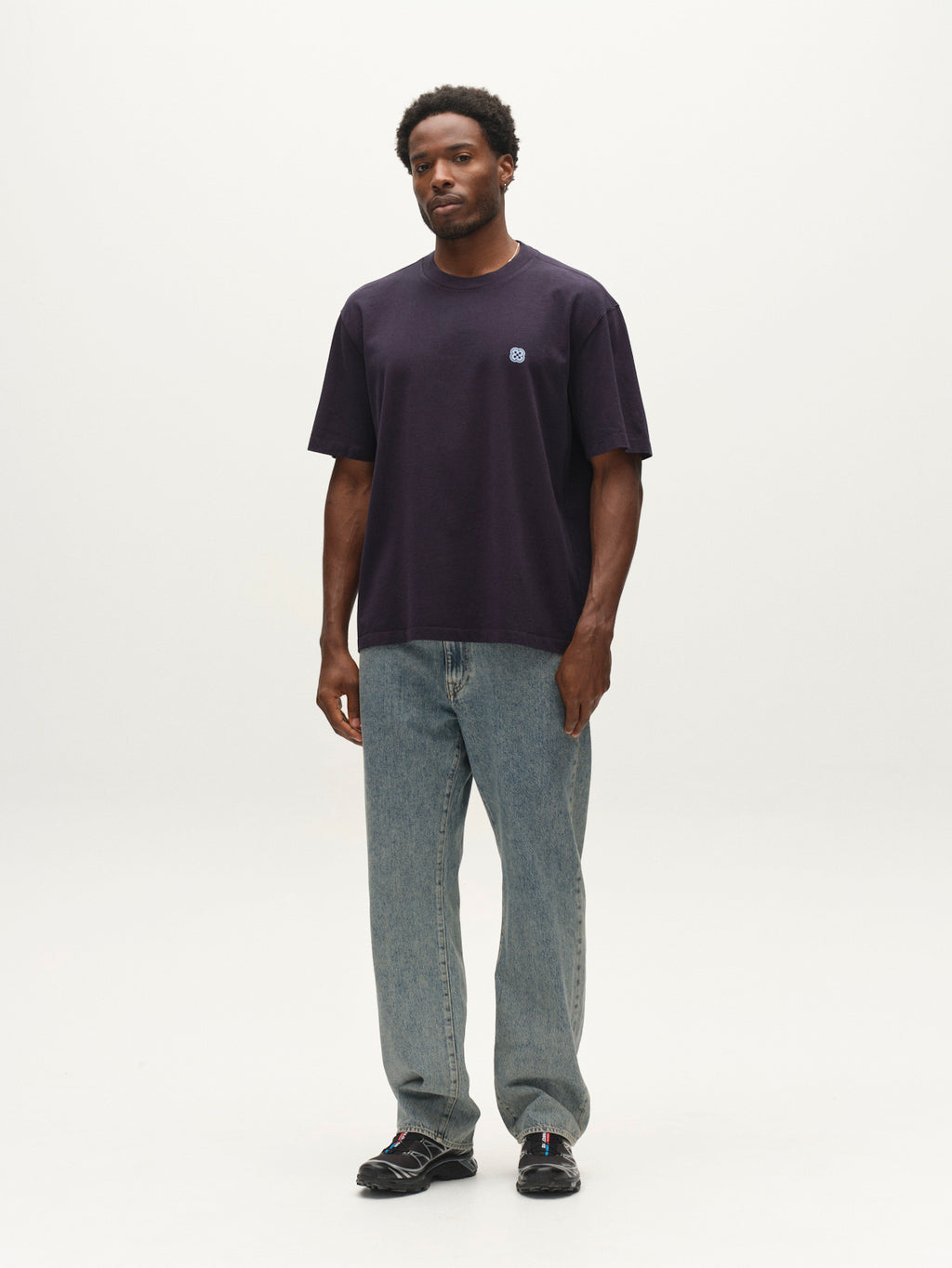 A man stands against a plain white background wearing the Gabba GABlegin Wix Boxy Hour Lobby2 SS T-shirt in Night Sky, paired with light blue jeans and black sneakers. He has short hair and a beard, with his arms relaxed by his sides. The T-shirt features a relaxed boxy fit for added comfort.