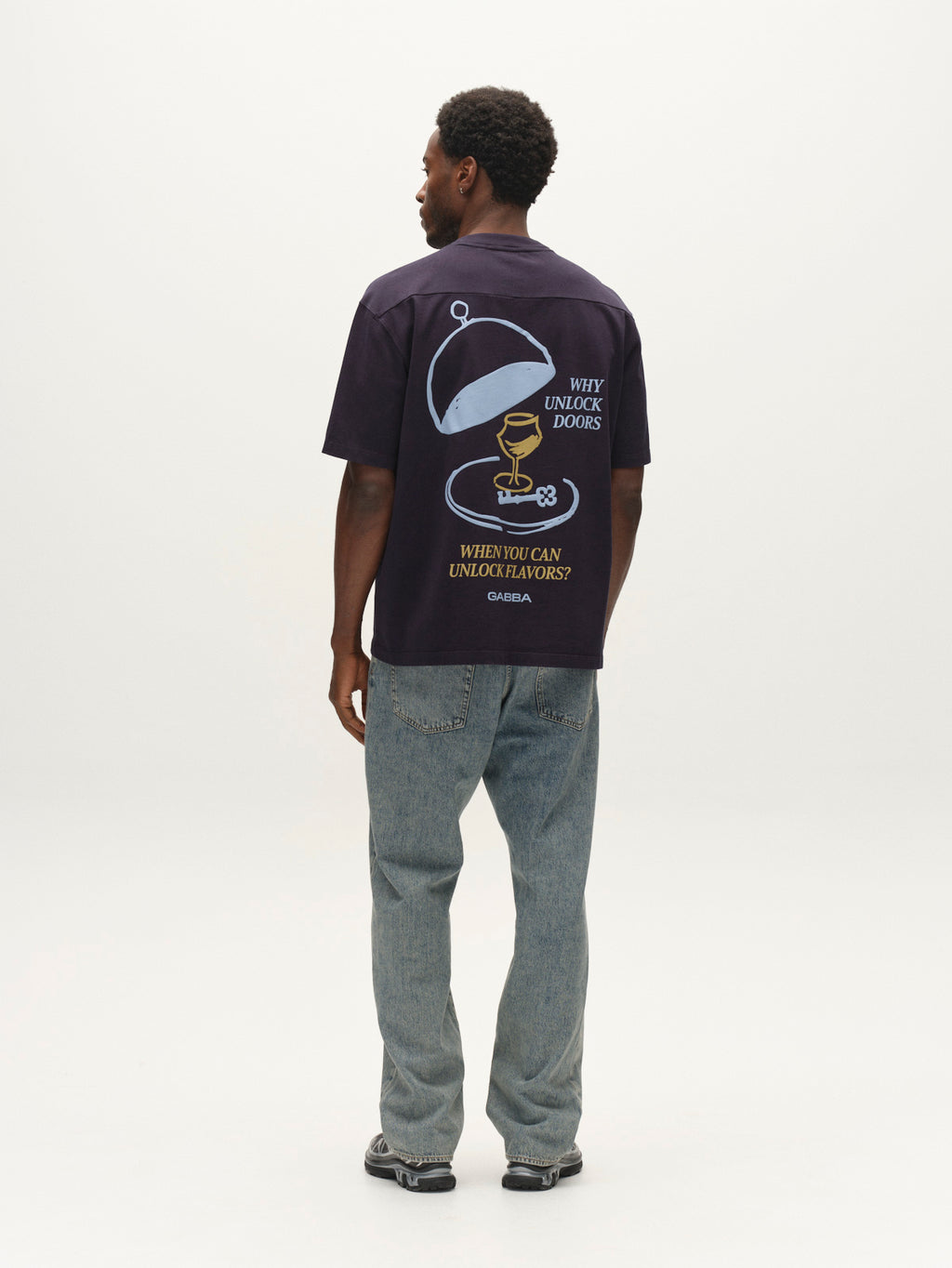 A man stands facing away, wearing the Gabba GABlegin Wix Boxy Hour Lobby2 SS T-shirt in Night Sky, crafted from textured cotton jersey. The black T-shirt features a graphic back print of a key, a dome lid, and the text “Why unlock doors when you can unlock flavors? Omsom.” Its relaxed boxy fit pairs effortlessly with light blue jeans and sneakers.