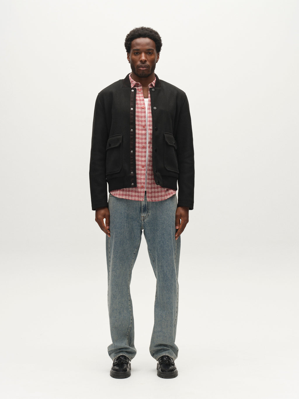 A man stands facing forward, wearing the Gabba GABmatheo Plain Bomber Jacket in black over a red plaid shirt, blue jeans, and black shoes, against a plain white background.