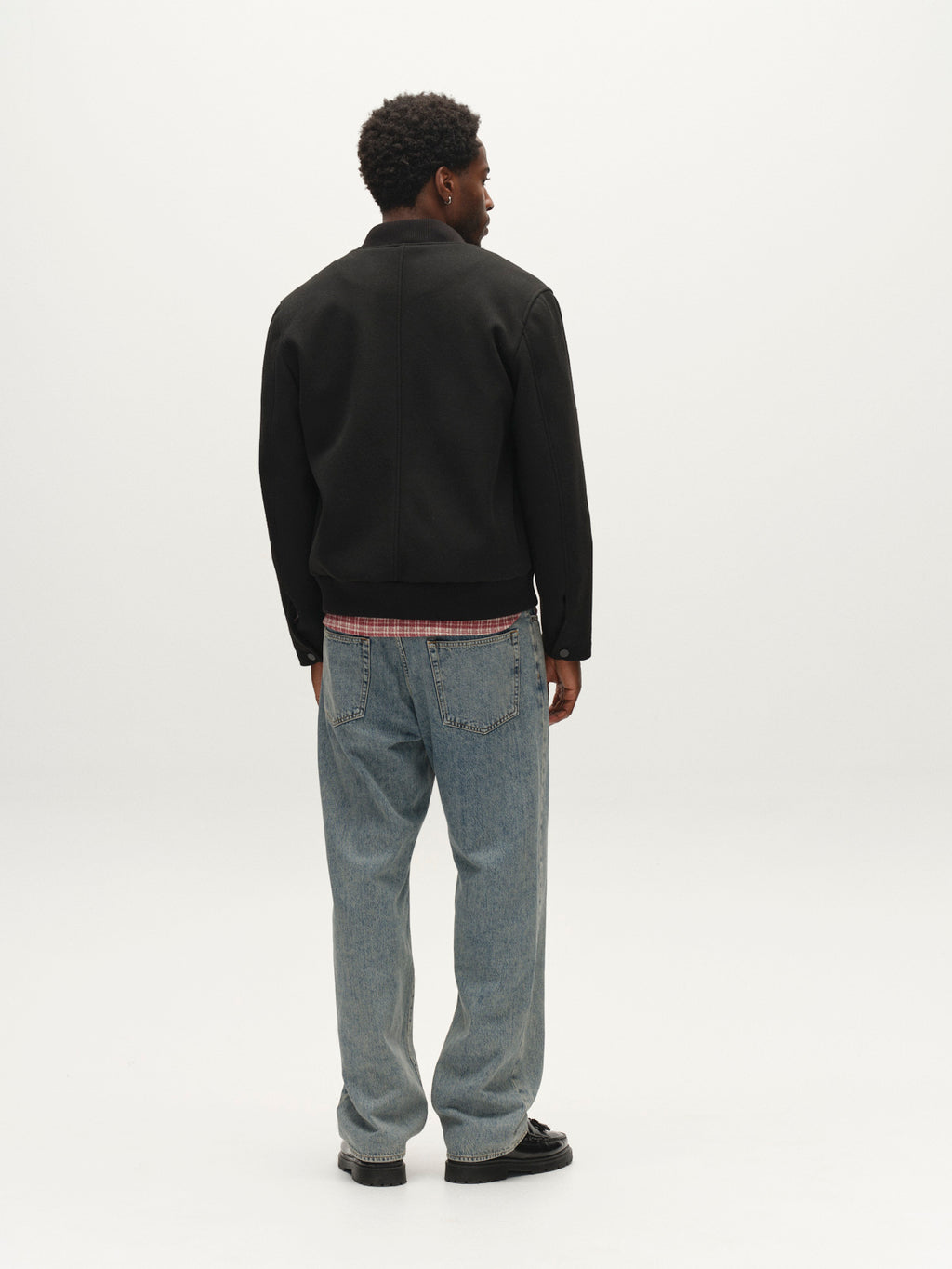 A man stands facing away, wearing the Gabba GABmatheo Plain Bomber Jacket in black over a plaid shirt, paired with light blue jeans and black shoes against a plain white background.