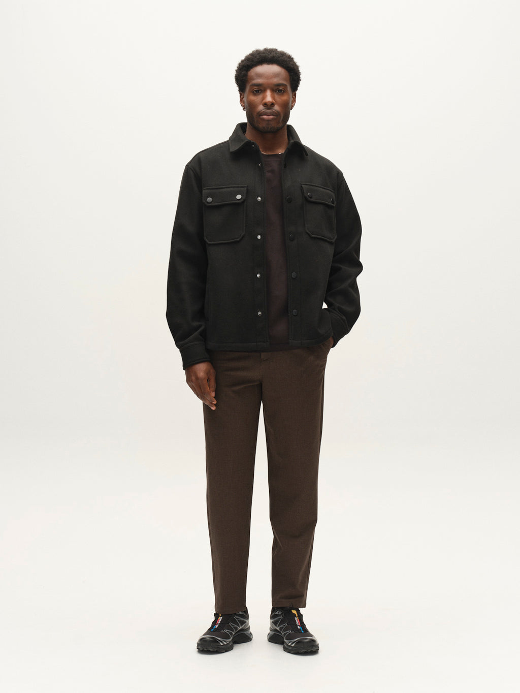 A man stands facing forward wearing the Gabba GABtheo City Jacket in black, paired with brown pants, a dark shirt, and black sneakers against a plain, light background—an ideal look for everyday layering.
