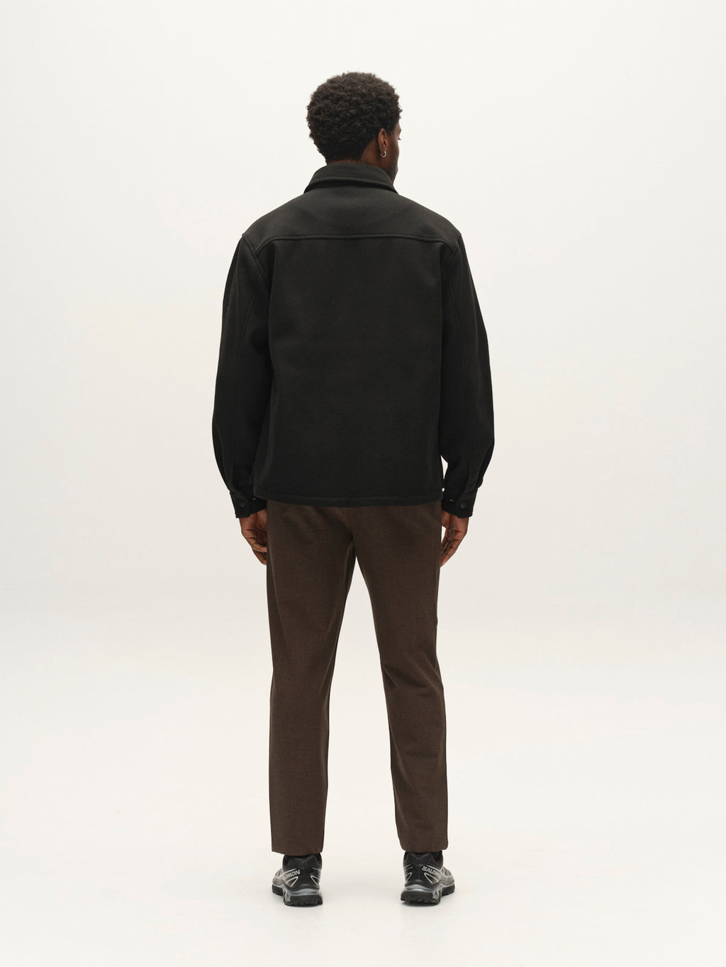 A man stands facing away, wearing the Gabba GABtheo City Jacket in black, paired with brown pants and black shoes against a plain, light background—effortlessly styled for everyday layering.