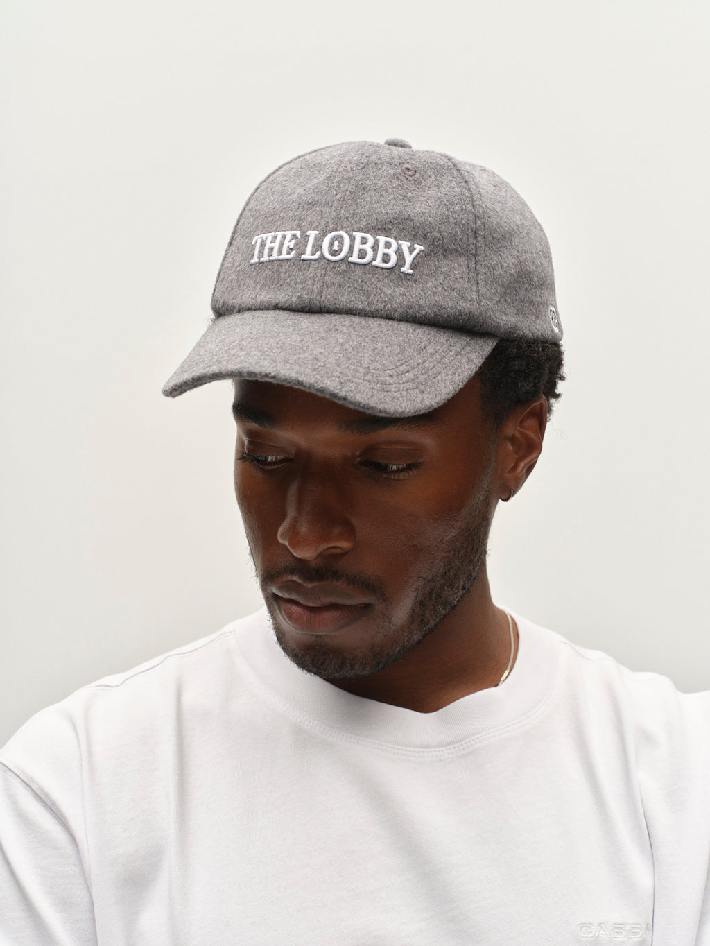 A man wearing the Gabba GABlimo W Lobby Cap in Grey Mel, featuring “THE LOBBY” embroidered in white letters, and a white shirt looks down against a plain light background.