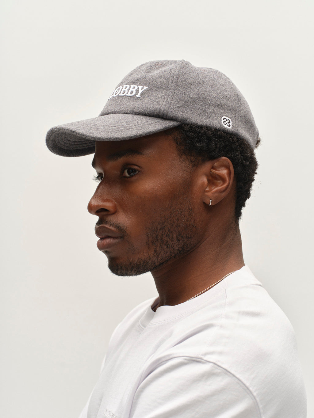 A man wearing the Gabba GABlimo W Lobby Cap in Grey Mel and a white shirt is shown in profile against a plain white background. He has a short beard, an earring, and looks ahead with a neutral expression.