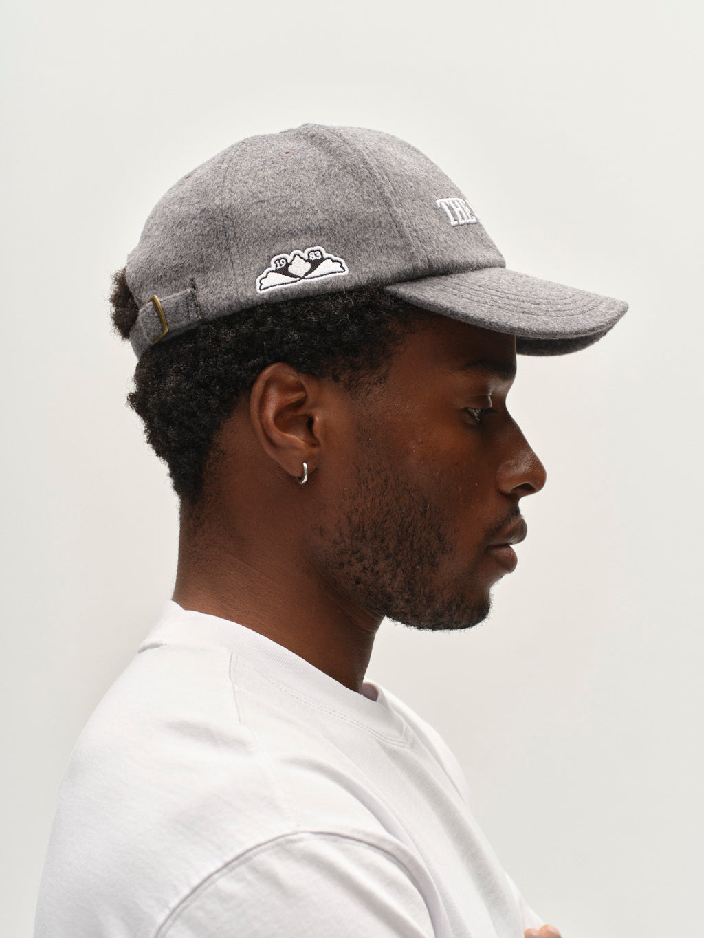 A person is shown in profile against a plain light background, wearing a white shirt and the Gabba GABlimo W Lobby Cap in Grey Mel. They have short curly hair and wear a small hoop earring.