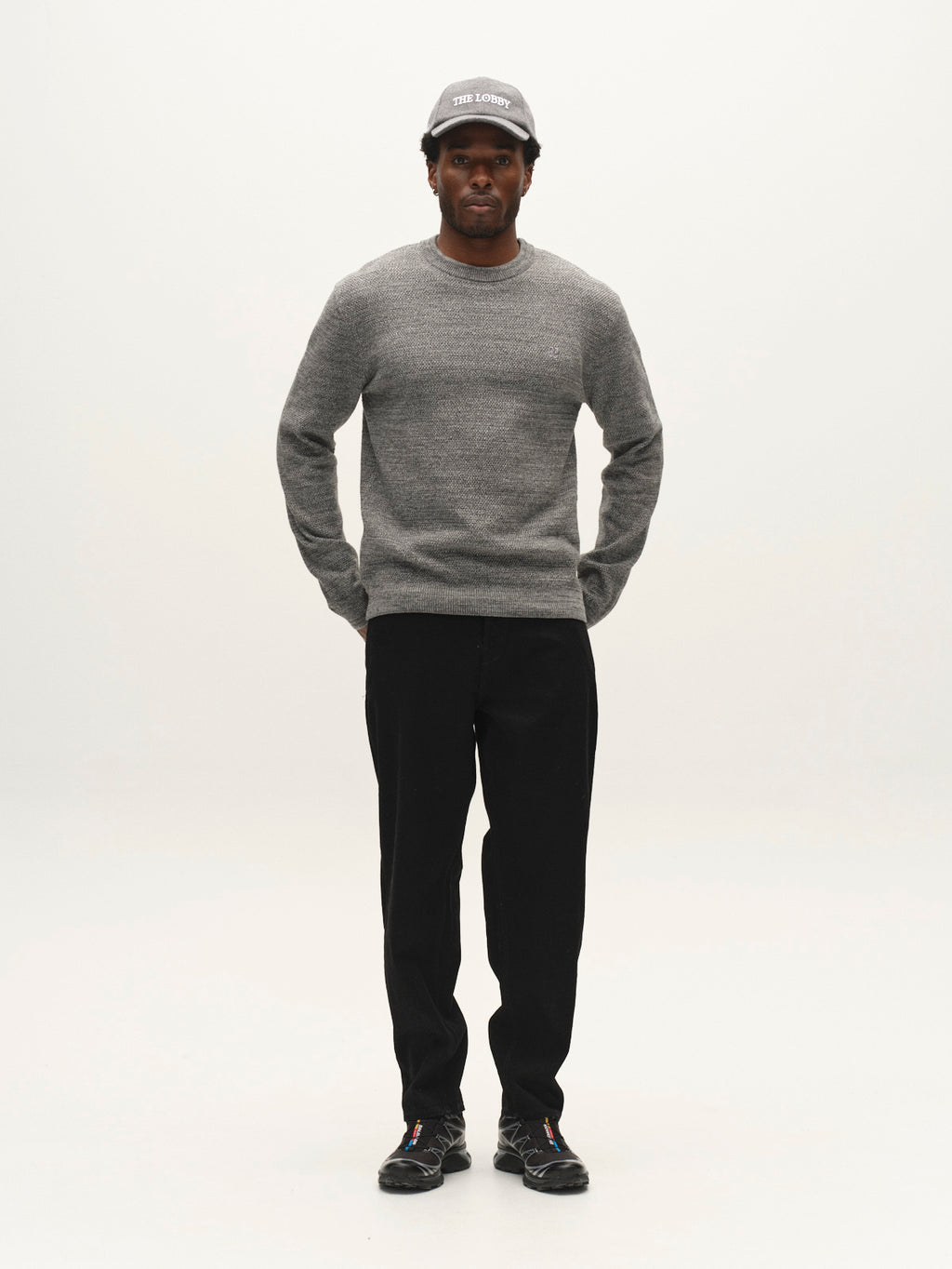 A man stands facing forward, wearing the Gabba GABlamix Coro O-Neck LS Knit sweater in light grey, paired with black pants, a gray cap, and black shoes, against a plain white background.