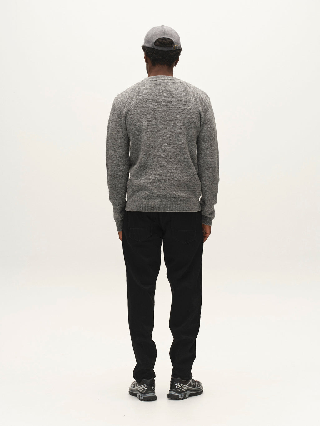 A person stands facing away, wearing the GABlamix Coro O-Neck LS Knit in Light Grey from Gabba, paired with black pants, gray sneakers, and a gray cap. The background is plain white.