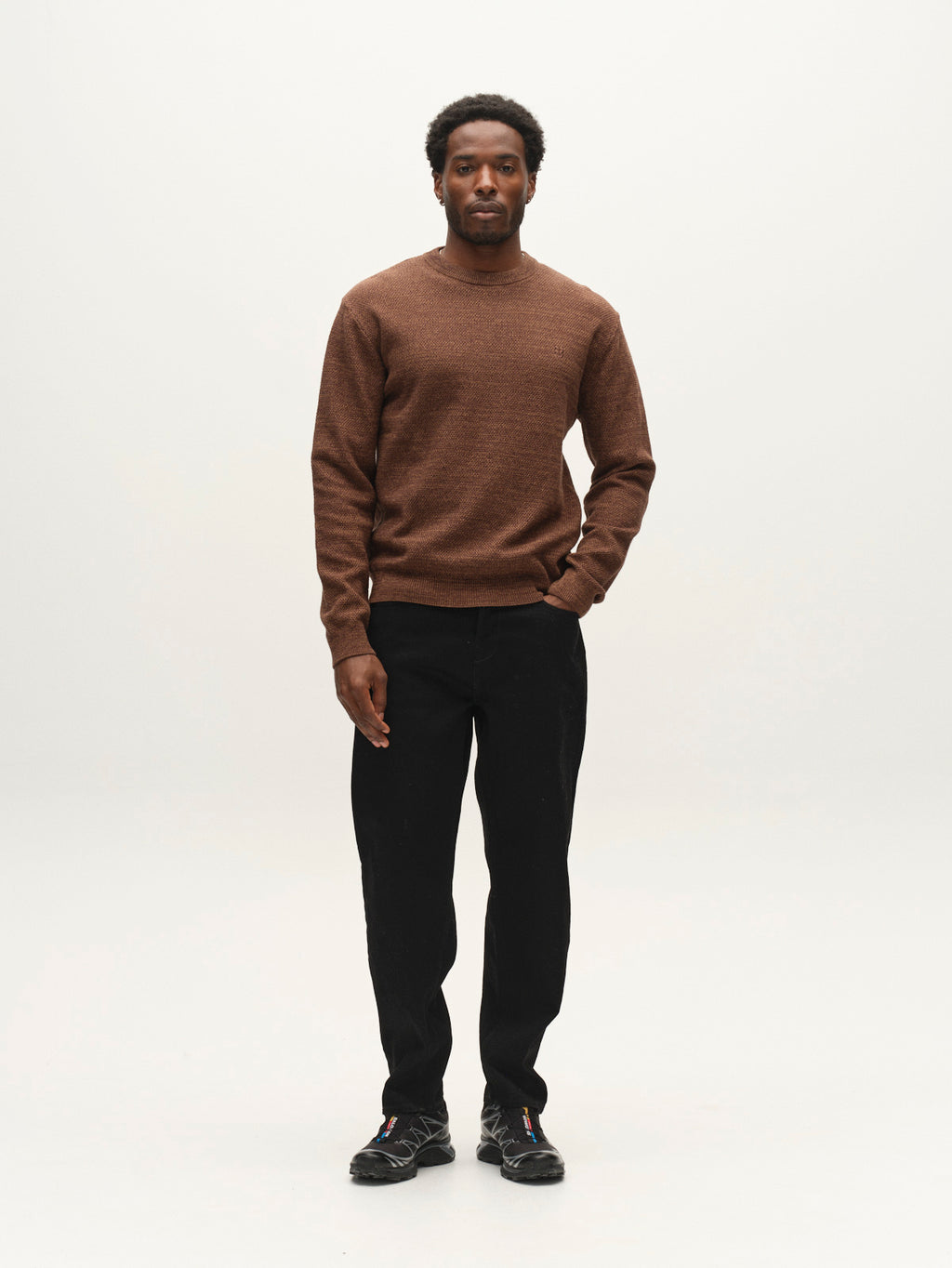 A person stands facing forward, wearing the Gabba GABlamix Coro O-Neck LS Knit in Brown Slate, paired with black pants and black shoes, against a plain white background.