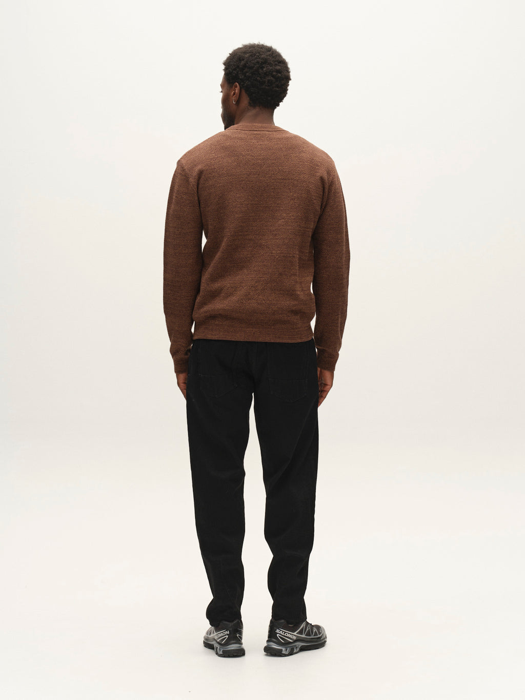 A man stands facing away from the camera, wearing the Gabba GABlamix Coro O-Neck LS Knit in Brown Slate, paired with black pants and black shoes, against a plain, light background.