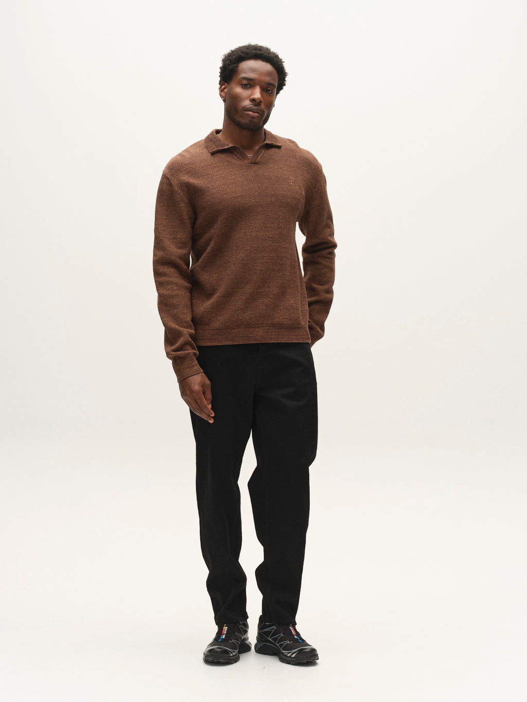 A man stands against a plain white background, wearing the Gabba GABlamix Coro Jhon LS Knit in Brown Slate—a cotton-blend knit long-sleeve shirt with a ribbed polo collar—paired with black pants and black athletic shoes. He has short curly hair and a neutral expression, with one hand in his pocket.