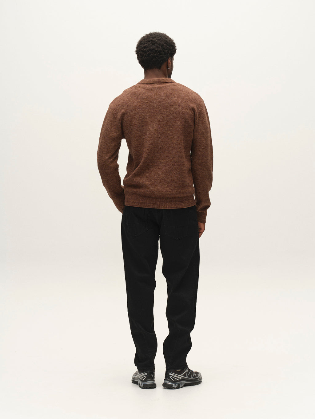 A person with short curly hair, wearing the Gabba GABlamix Coro Jhon LS Knit in Brown Slate, black pants, and gray sneakers stands facing away against a plain, light background.