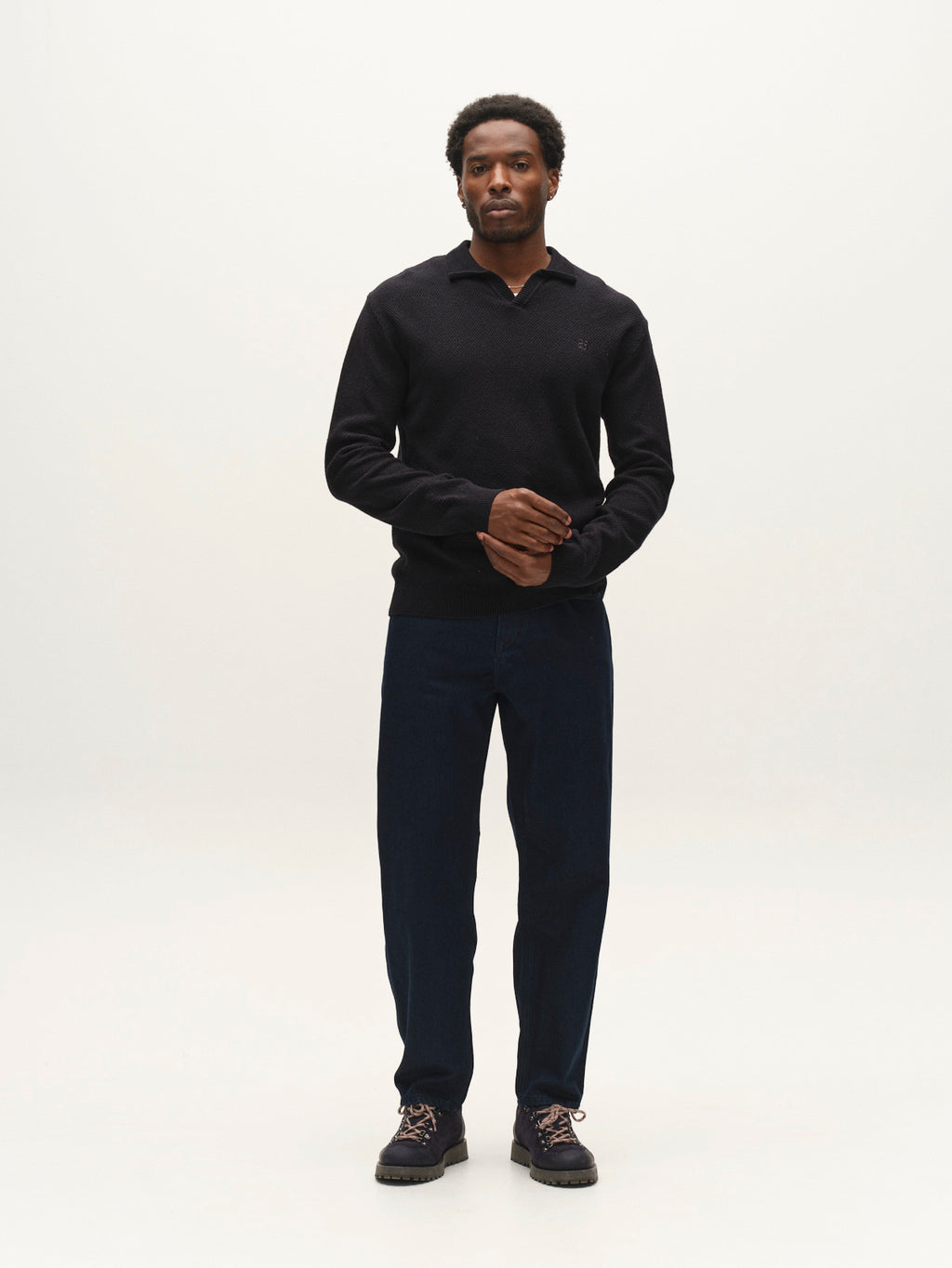 A man stands against a plain white background, wearing the Gabba GABlamix Coro Jhon LS Knit in Blue Black—a refined black long-sleeve shirt with a ribbed polo collar and elegant knit—paired with dark blue pants and dark shoes. He faces forward with his hands loosely clasped.