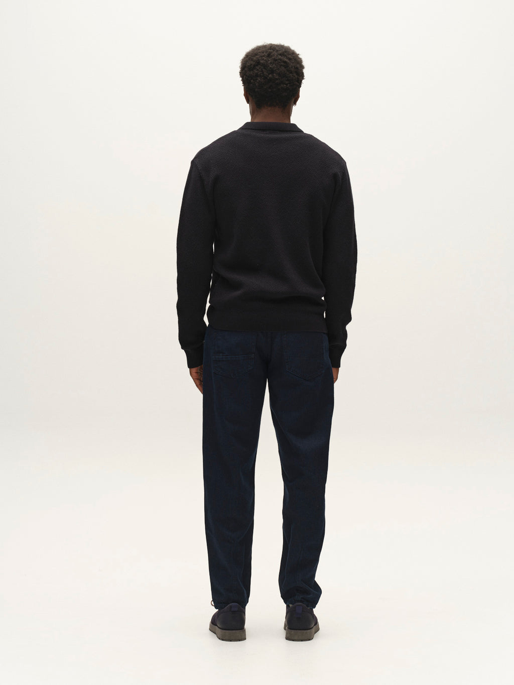 A person with short curly hair stands facing away from the camera, wearing the Gabba GABlamix Coro Jhon LS Knit in Blue Black, paired with dark blue pants and dark shoes, set against a plain light background.