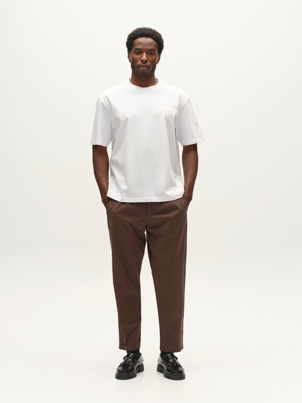 A man stands facing forward against a plain white background, wearing the Gabba GABlegin Wix Boxy Hour SS T-shirt in white, brown pants, and black shoes.