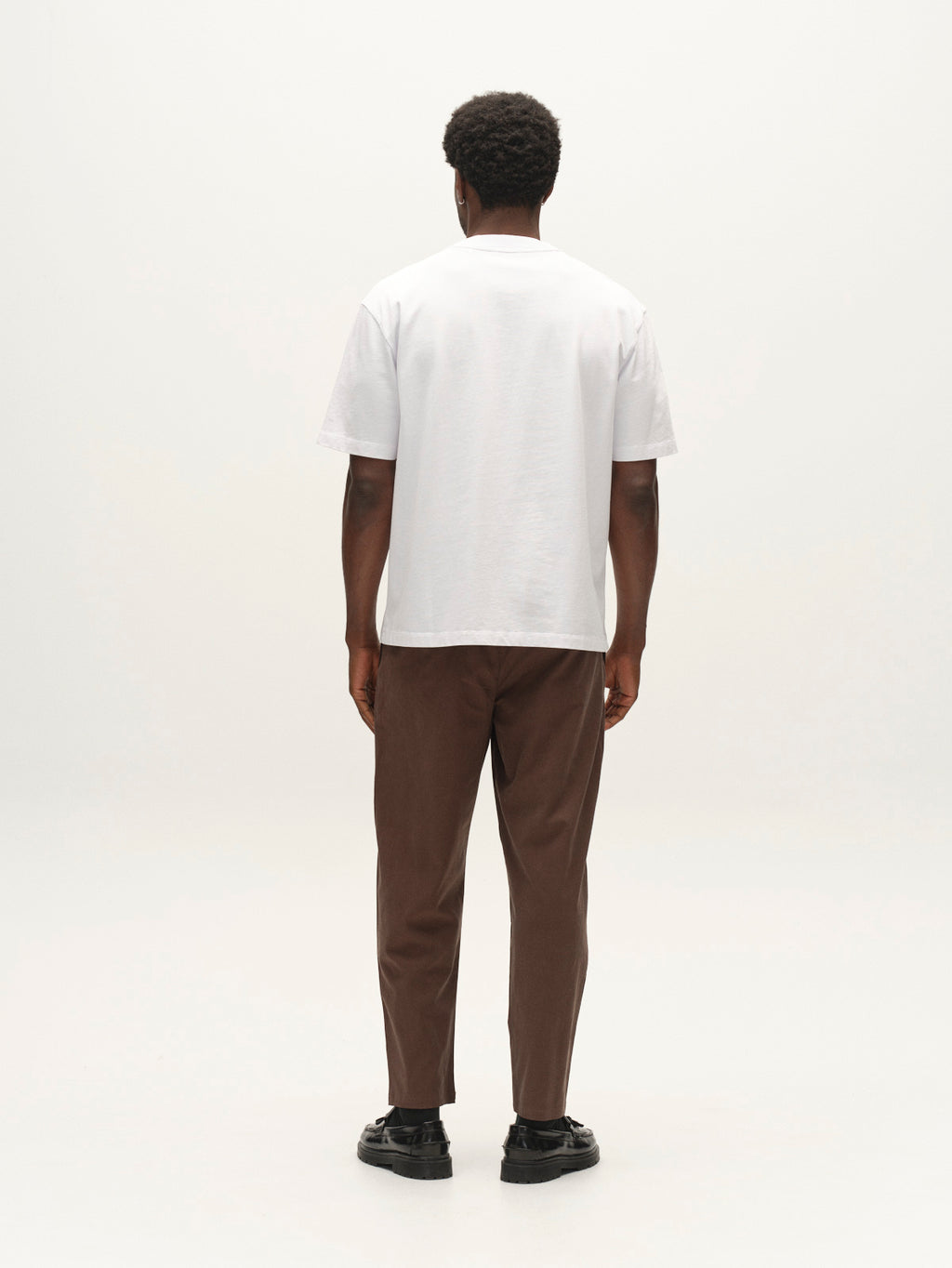 A person stands facing away from the camera, wearing the Gabba GABlegin Wix Boxy Hour SS T-shirt in white—a relaxed, boxy-fit tee crafted from soft cotton jersey—paired with brown pants and black shoes against a white background. The embroidered Gabba logo offers a subtle accent on the back of the t-shirt.
