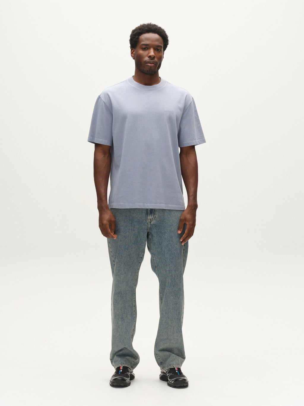 A man stands facing forward, wearing the Gabba GABlegin Wix Boxy Hour SS T-shirt in Dusty Blue. The t-shirt has a relaxed boxy fit and is crafted from garment-washed cotton, featuring an embroidered Gabba logo for a subtle yet distinctive touch. He pairs it with blue jeans and black shoes against a plain white background.
