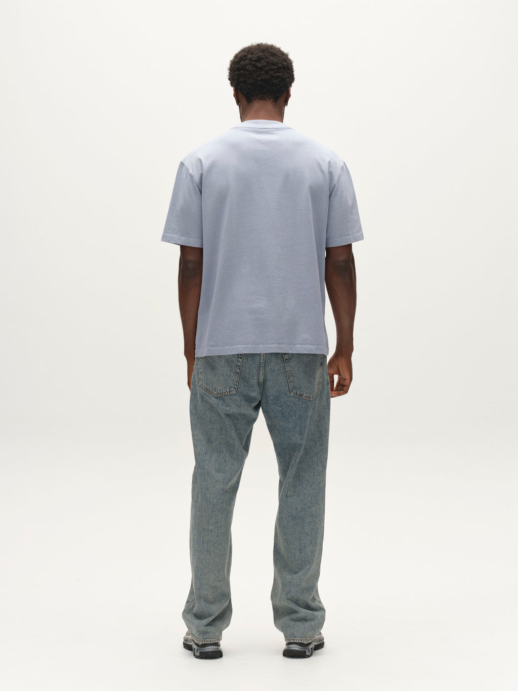 A person stands facing away from the camera, wearing the Gabba GABlegin Wix Boxy Hour SS T-shirt in Dusty Blue—a garment-washed cotton piece designed with a relaxed boxy fit—paired with loose-fitting blue jeans and black shoes, set against a plain white background.