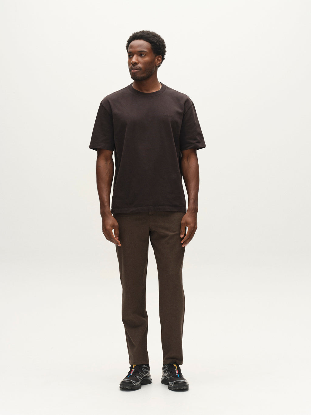 A man stands facing slightly to the side, wearing the Gabba GABlegin Wix Boxy Hour SS T-shirt in black—a mid-weight cotton jersey tee with an embroidered Gabba logo—paired with brown pants and black shoes, against a plain light background. The T-shirt features a relaxed boxy fit, lending a casual yet refined appearance.