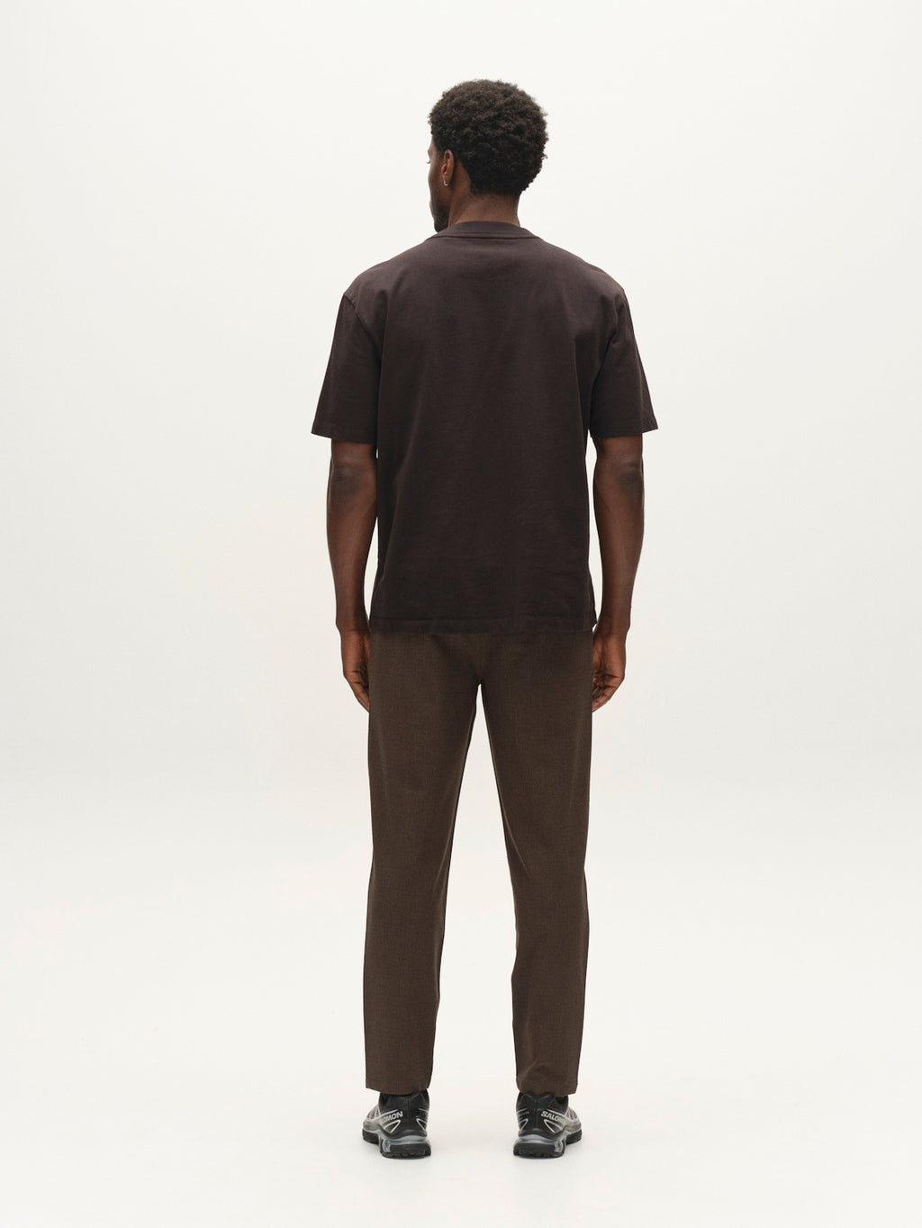 A man stands facing away from the camera, wearing the Gabba GABlegin Wix Boxy Hour SS T-shirt in black, paired with brown pants and black shoes, against a plain white background.