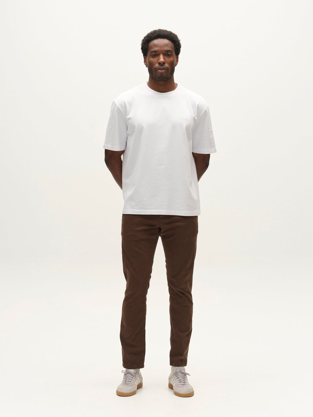 A man stands facing forward with his hands behind his back, wearing a plain white T-shirt, the Gabba GABpaul K3280 Dale Chino in Brown Slate—crafted from high-stretch cotton-blend fabric—and white sneakers against a plain white background, showcasing a timeless look in everyday wear.