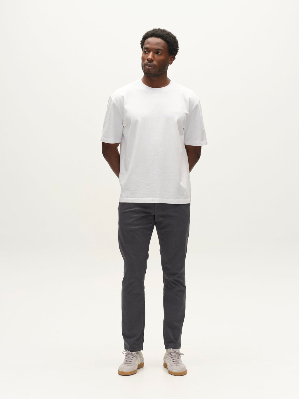 A man stands against a plain white background, wearing a white t-shirt, light sneakers, and Gabba GABpaul K3280 Dale Chino pants in Turbulence Blue with French pockets and double-paspel back pockets. His hands are behind his back as he looks slightly to the side.