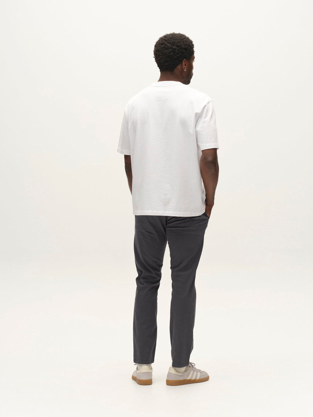 A man stands facing away, wearing a white T-shirt, Gabba GABpaul K3280 Dale Chino in Turbulence Blue with double-paspel and French pockets, and beige sneakers, his hands in his pockets against a plain light background.