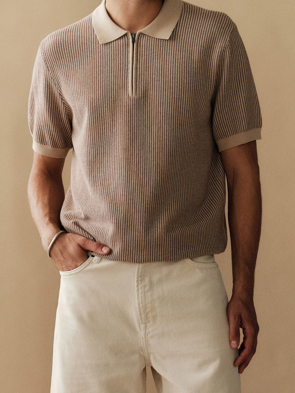 A man wears the Gabba GABoswald Retro SS Knit in Walnut, a short-sleeved knit polo with 3D rib cotton and zip collar, paired with light beige pants, standing against a plain beige background with one hand in his pocket.