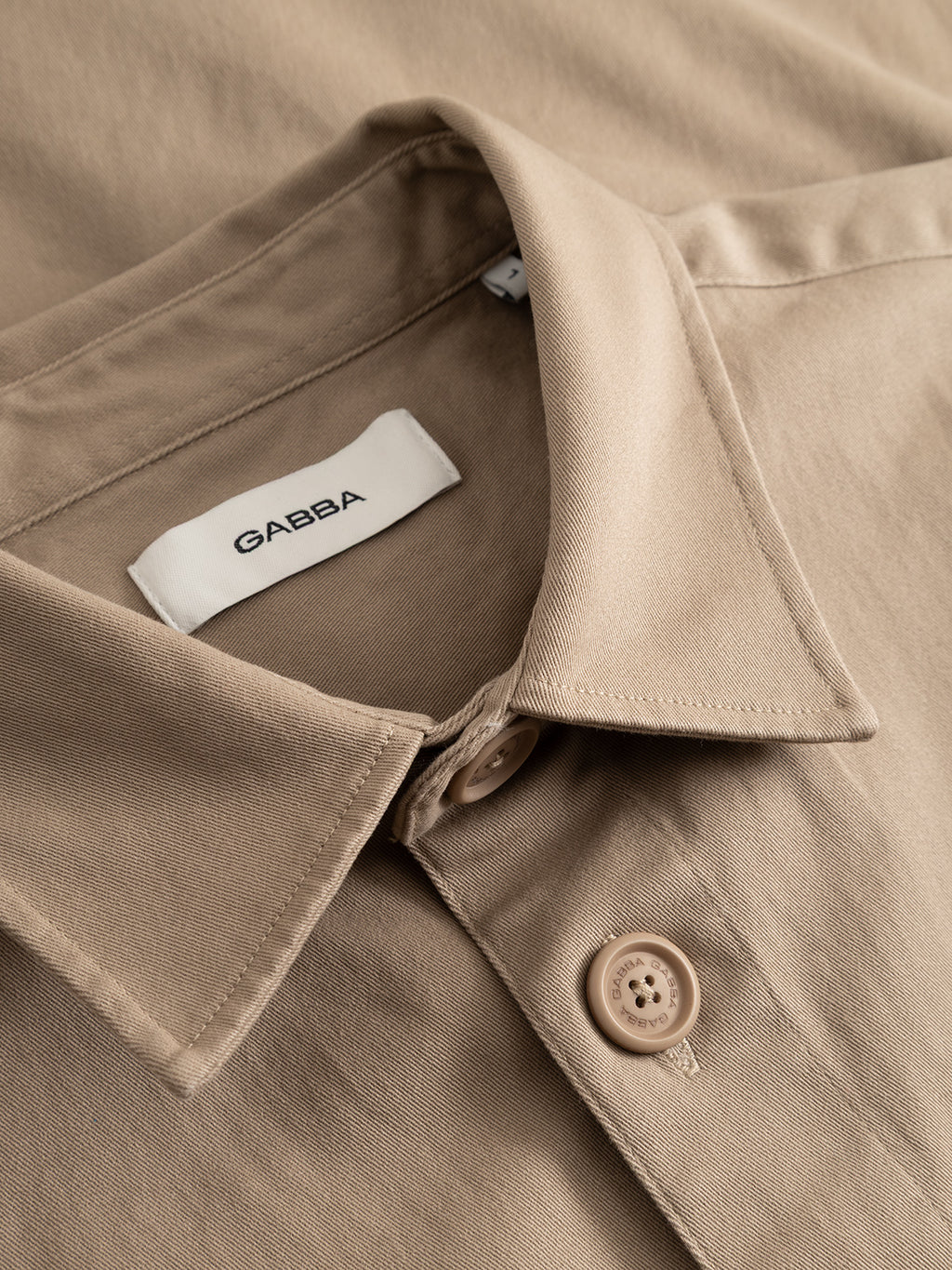 Close-up view of the GABtopper LS Shirt - Sand Plaza by Gabba, a beige collared design featuring button details and a visible white Gabba label inside the collar. The shirt is neatly folded, showcasing its textured fabric.