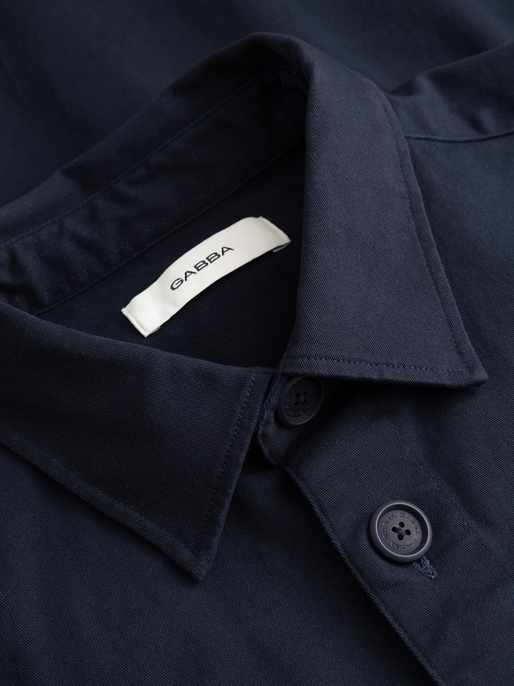 Close-up of the GABtopper LS Shirt in Blue Black from Gabba, featuring a navy blue collar with two buttons visible and a white label inside the collar displaying the brand name. This regular fit shirt is an essential layering piece, perfect for adding effortless style to your everyday wardrobe.