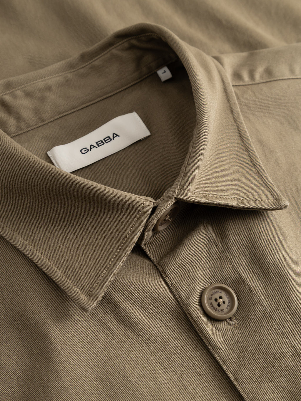 Close-up of the GABtopper LS Shirt in Dusty Olive, featuring a beige collared design with buttons. A white label with the brand name Gabba is sewn inside the collar. This heavyweight overshirt is crafted as a layering essential, with fabric that appears smooth and neatly pressed.