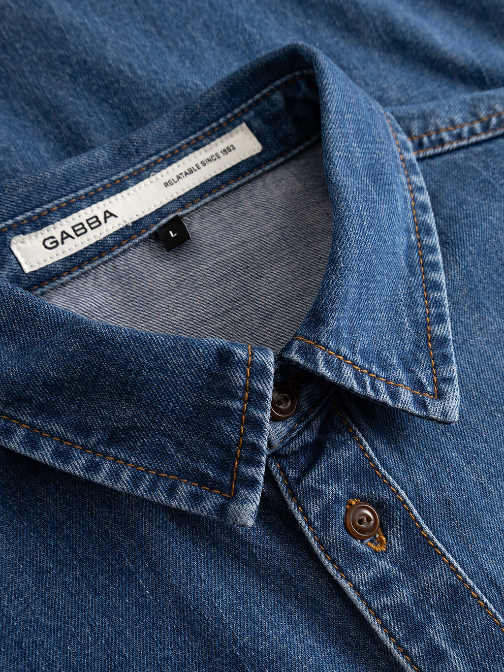 Close-up of the GABsven Reg Wish LS Shirt in mid blue denim, featuring a cotton collar with brown buttons and prominent orange stitching. A white label inside the collar displays the Gabba brand and size L, emphasizing this regular fit shirt as a versatile wardrobe essential.
