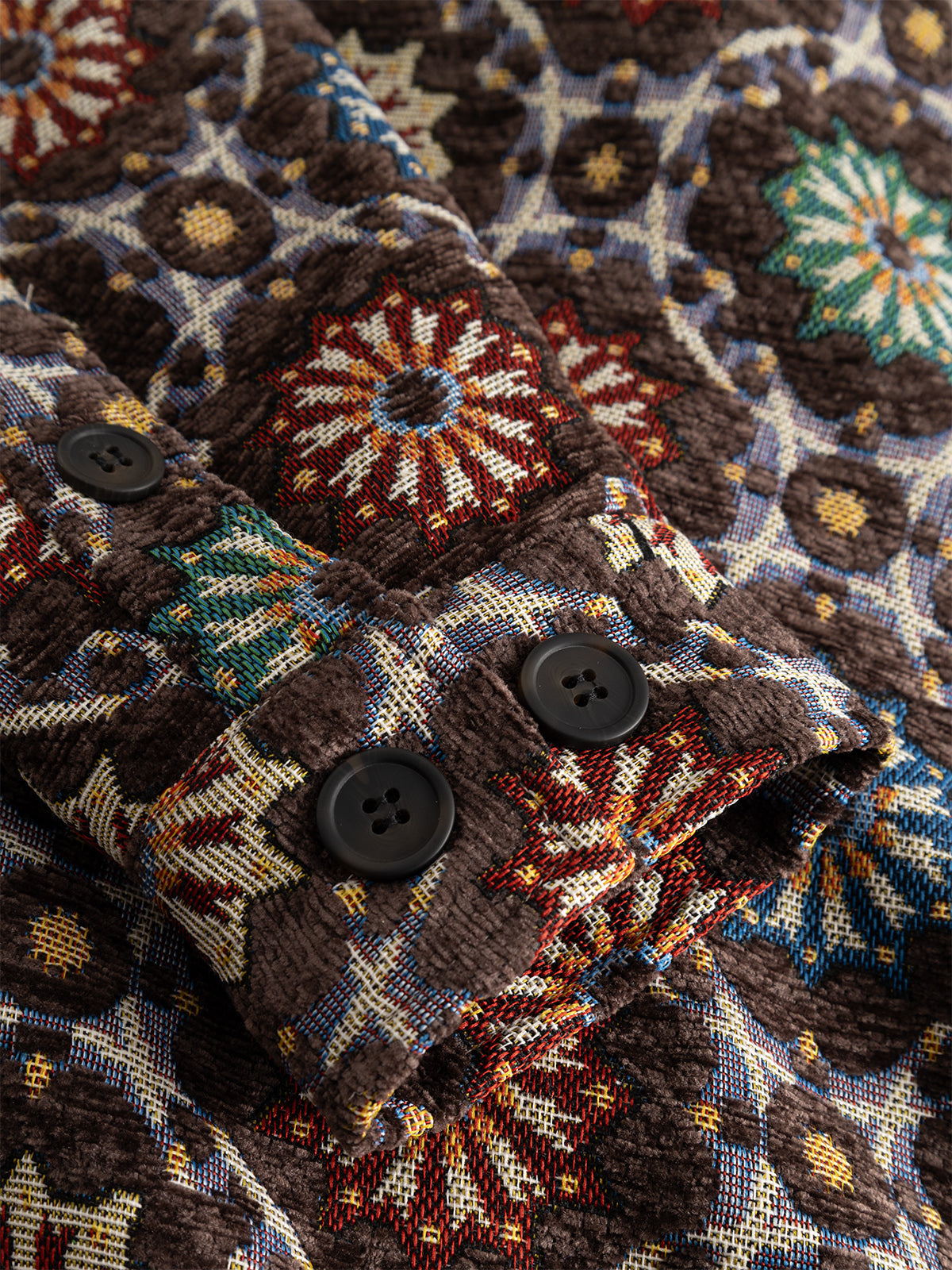 Close-up of the GABsimone Nava LS - Brown shirt by Gabba, showcasing a textured jacquard fabric with a vibrant geometric floral pattern. The design features red, green, white, and yellow accents on a brown background, along with two black buttons on the cuff, highlighting an abstract motif that adds visual interest to the garment.