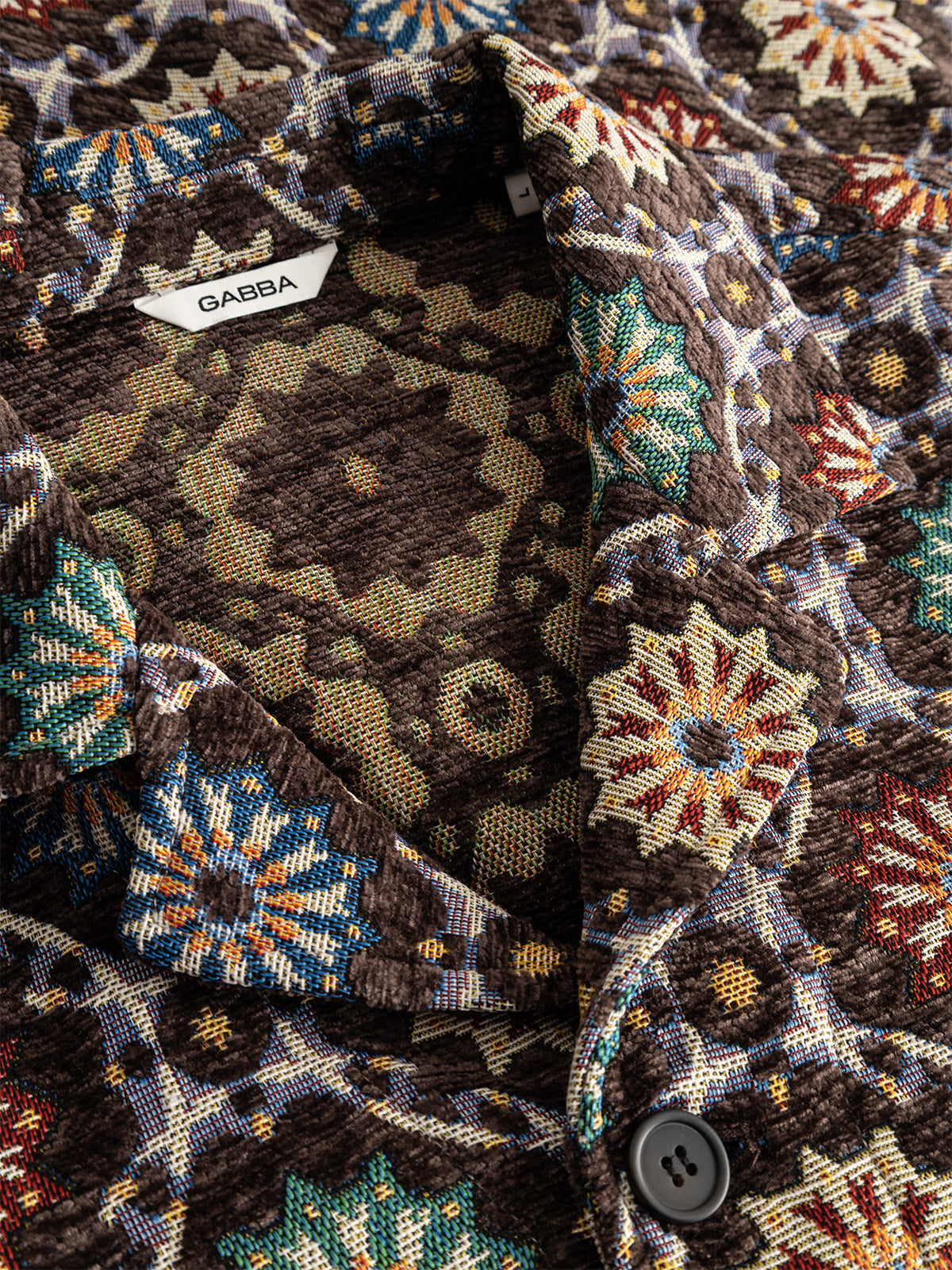 Close-up of the GABsimone Nava LS - Brown jacket by Gabba, crafted from textured jacquard fabric with a colorful geometric and abstract motif, showcasing a visible Gabba label, a resort collar, and a dark button near the neckline.