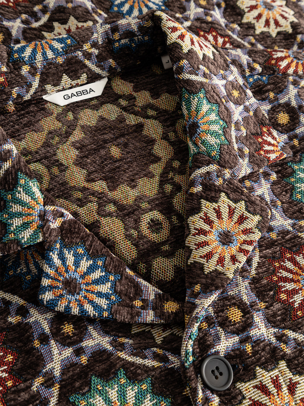 Close-up of the GABsimone Nava LS - Brown jacket by Gabba, crafted from textured jacquard fabric with a colorful geometric and abstract motif, showcasing a visible Gabba label, a resort collar, and a dark button near the neckline.