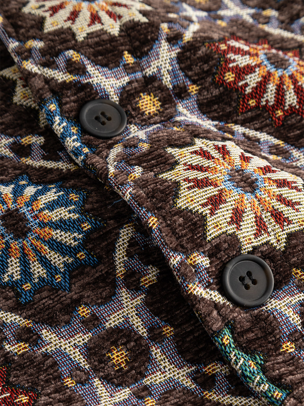 Close-up of the textured jacquard fabric on the Gabba GABsimone Nava LS - Brown, showcasing vibrant geometric star motifs in blue, yellow, red, and white. Two black buttons are attached to the brown material, suggesting garment detailing. The abstract pattern and button placement hint at features like a resort collar for a stylish touch.