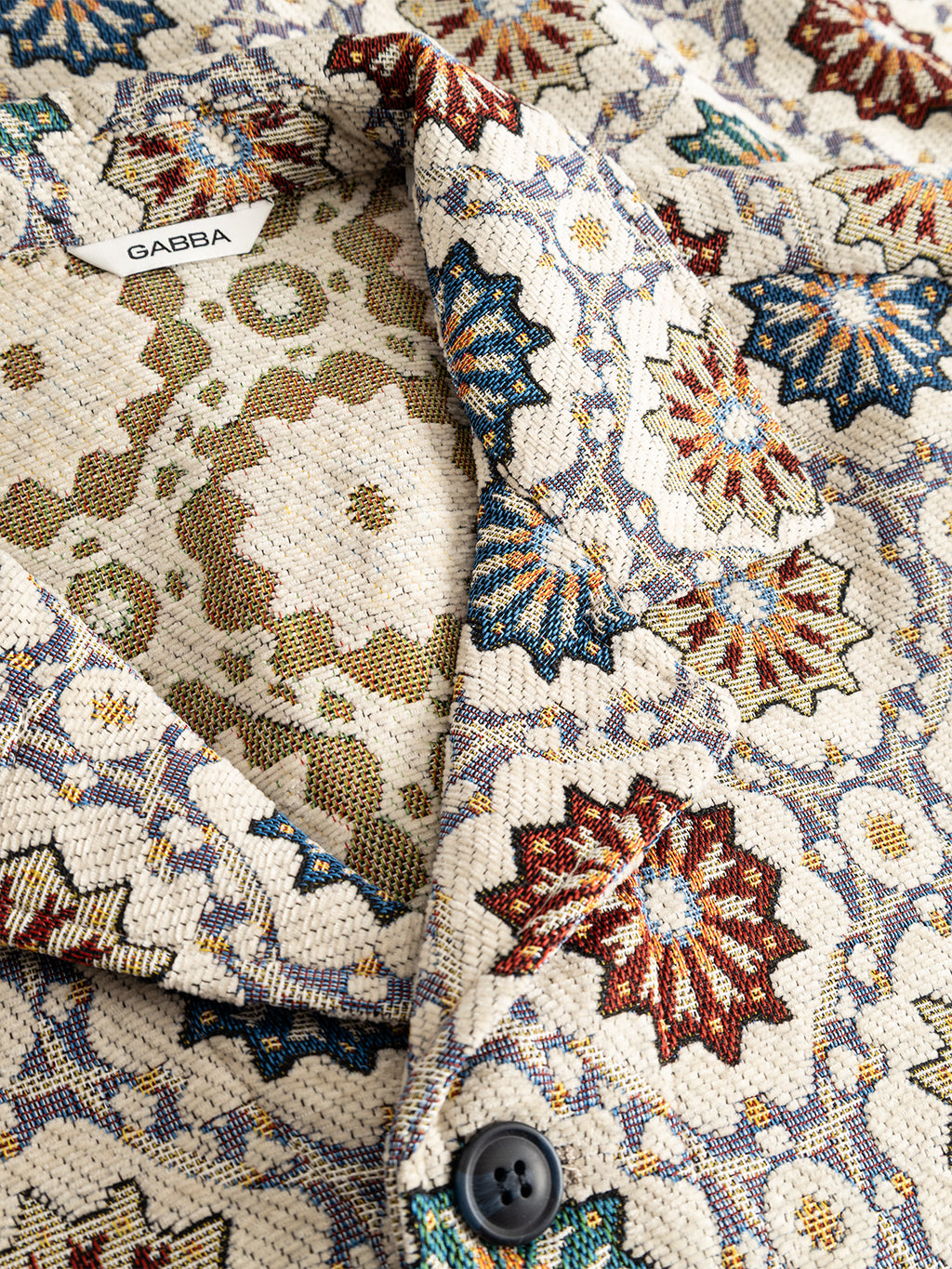 Close-up of the GABsimone Nava LS - Birch shirt by Gabba, crafted from patterned jacquard fabric featuring a geometric starburst design in red, blue, and beige tones. The shirt showcases a visible resort collar, button detail, and a white tag with the Gabba brand name.