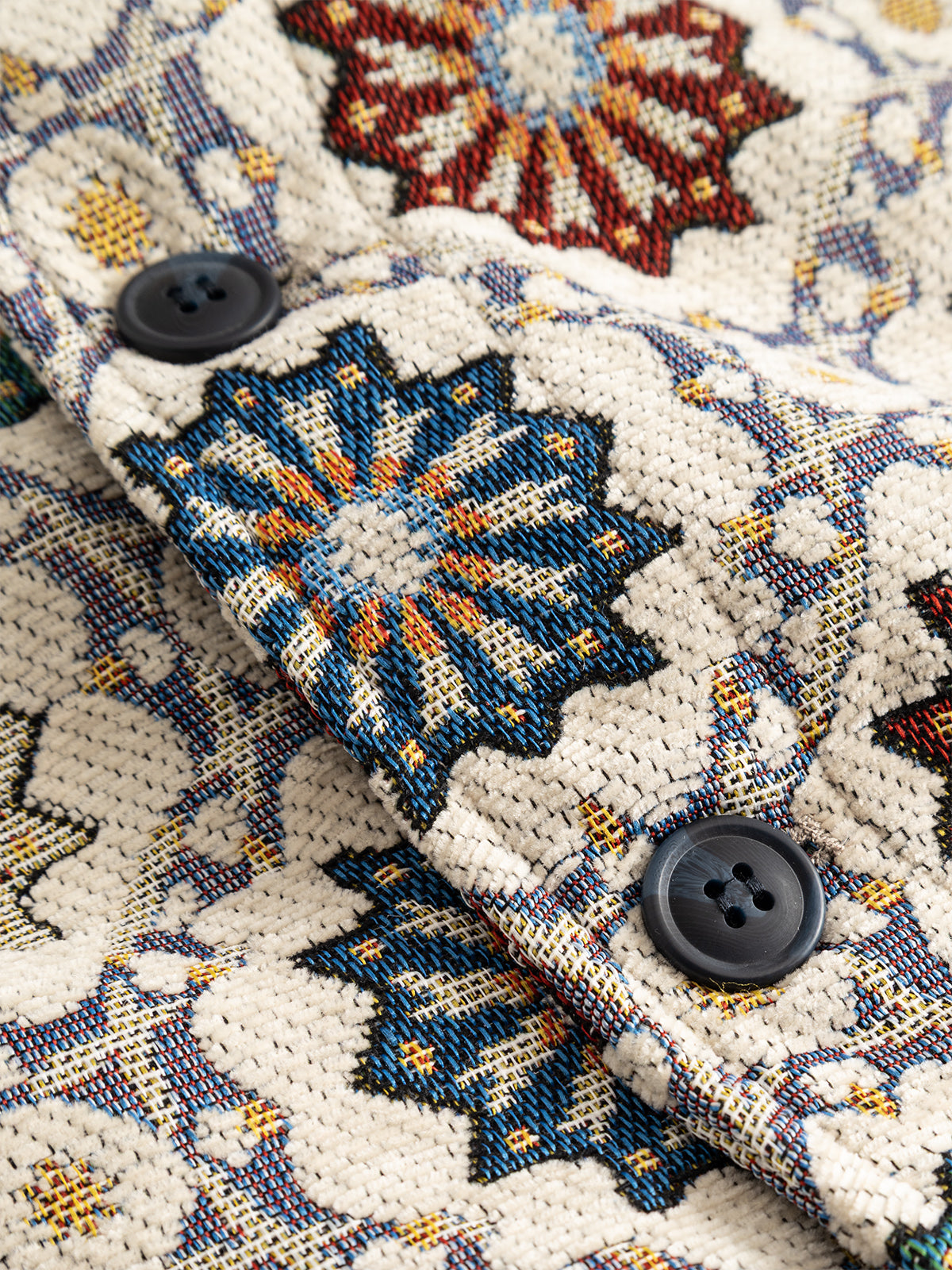 Close-up of the Gabba GABsimone Nava LS - Birch, showcasing its jacquard fabric featuring colorful geometric floral patterns in cream, blue, red, and yellow tones. Two black buttons are sewn onto the woven, embroidered-looking material.