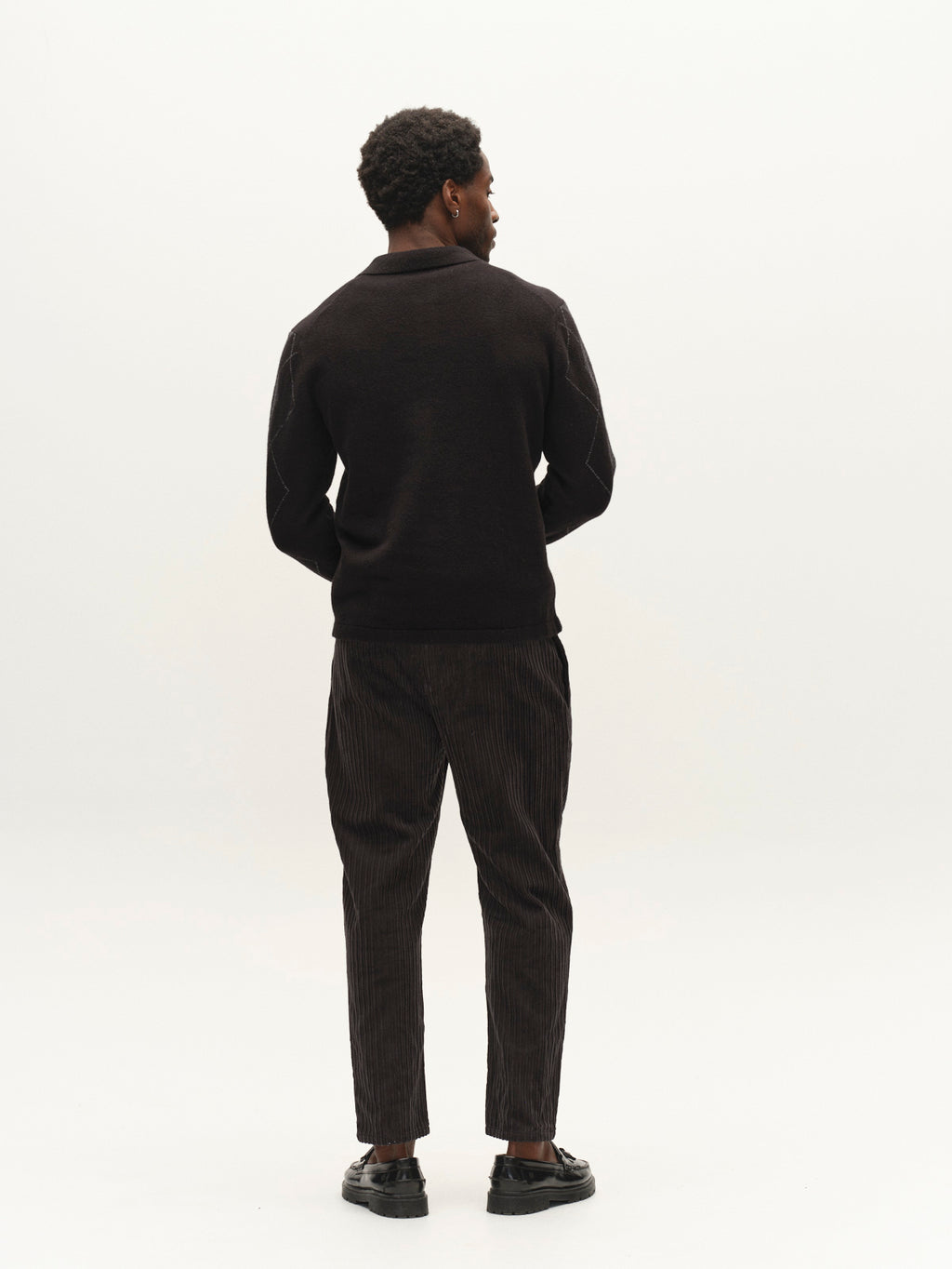 A person stands facing away from the camera, wearing the Gabba GABpietro Polo Argyle LS Knit in black, paired with dark pinstriped pants and black shoes, against a plain light background.