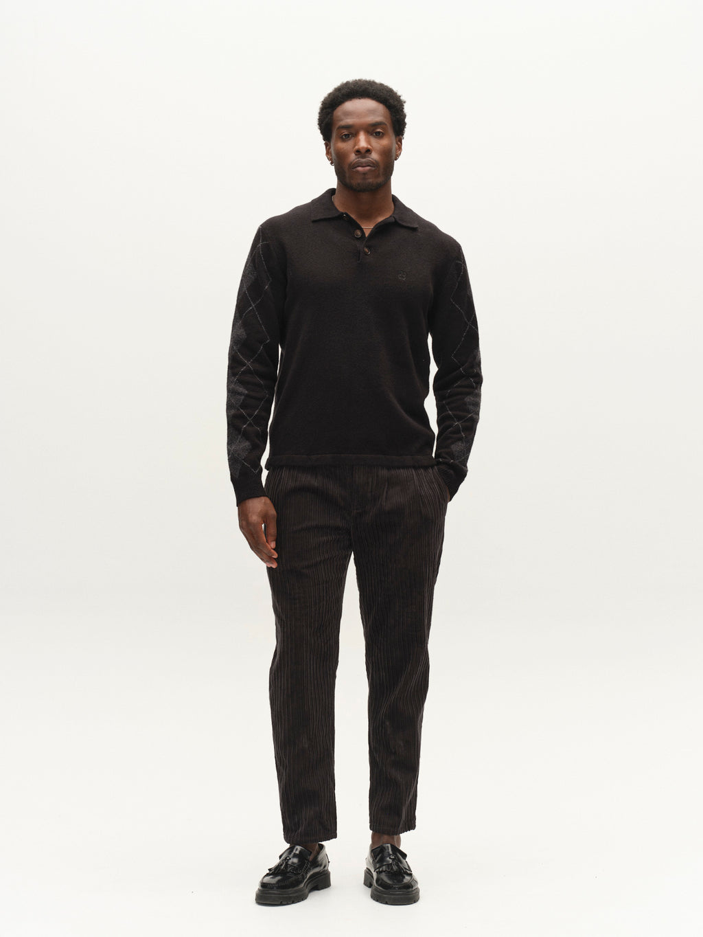 A man stands facing forward wearing the Gabba GABpietro Polo Argyle LS Knit in black, paired with brown striped pants and black shoes against a plain white background.