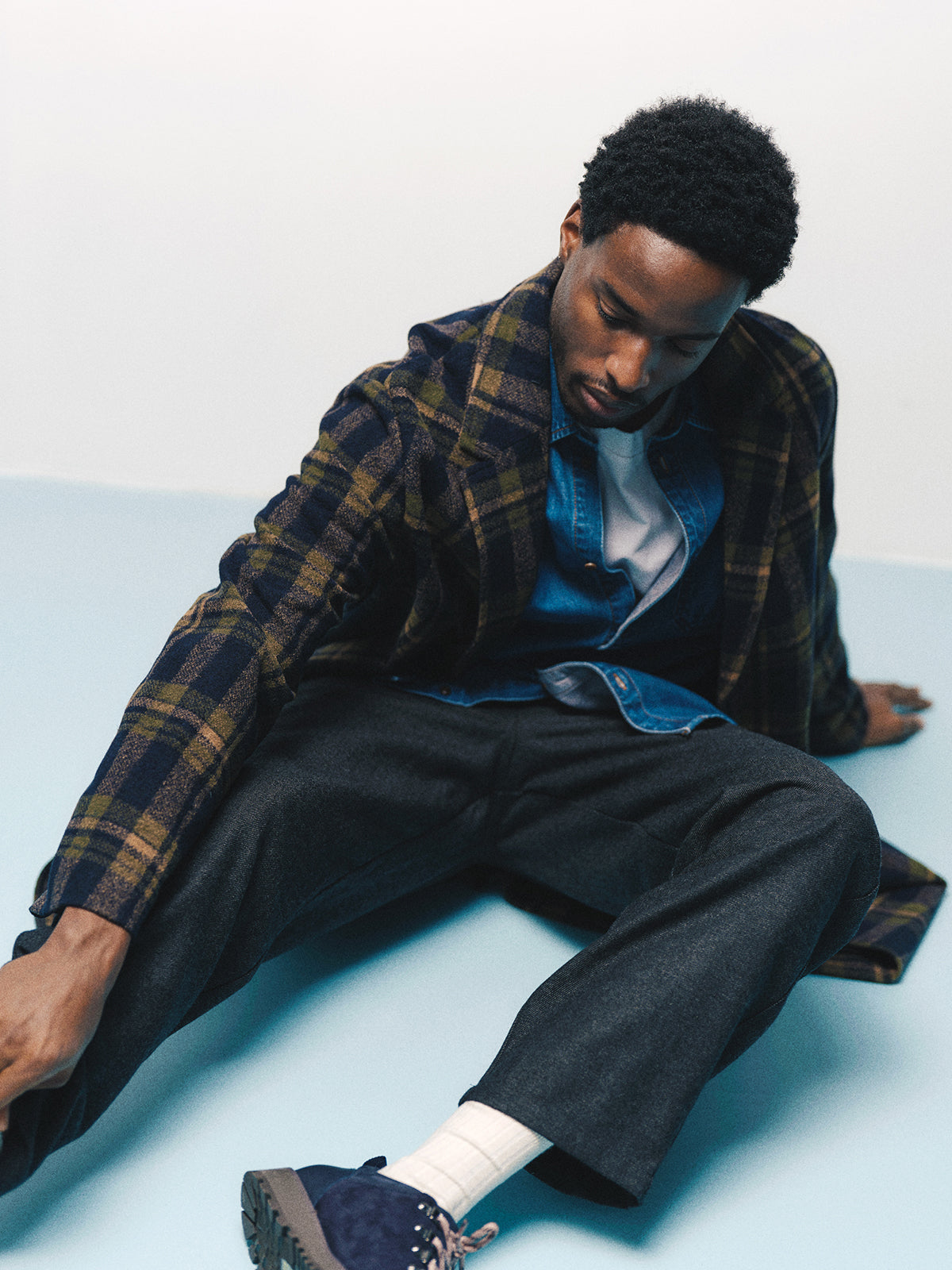 A man wearing a plaid coat, denim shirt, white t-shirt, and Gabba GABdavid Twilight Pant in Dark Grey with a loose tapered fit sits on the floor with one leg extended, looking down and reaching forward with his left arm.