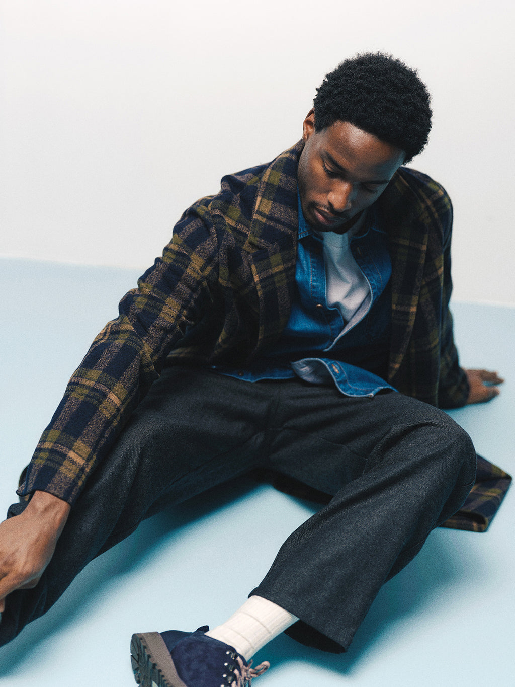 A man wearing a plaid coat, denim shirt, white t-shirt, and Gabba GABdavid Twilight Pant in Dark Grey with a loose tapered fit sits on the floor with one leg extended, looking down and reaching forward with his left arm.