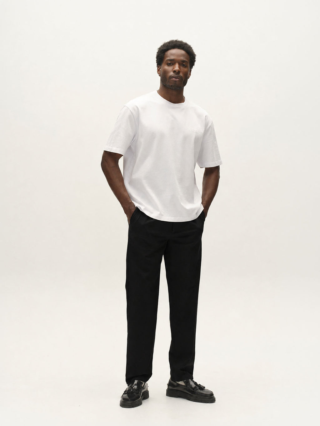 A man stands against a plain light background, wearing a white T-shirt, Gabba GABnapoli 19 Phin GRS - Black regular straight fit wool blend chino pants, and black shoes, with his hands in his pockets, looking at the camera with a neutral expression.