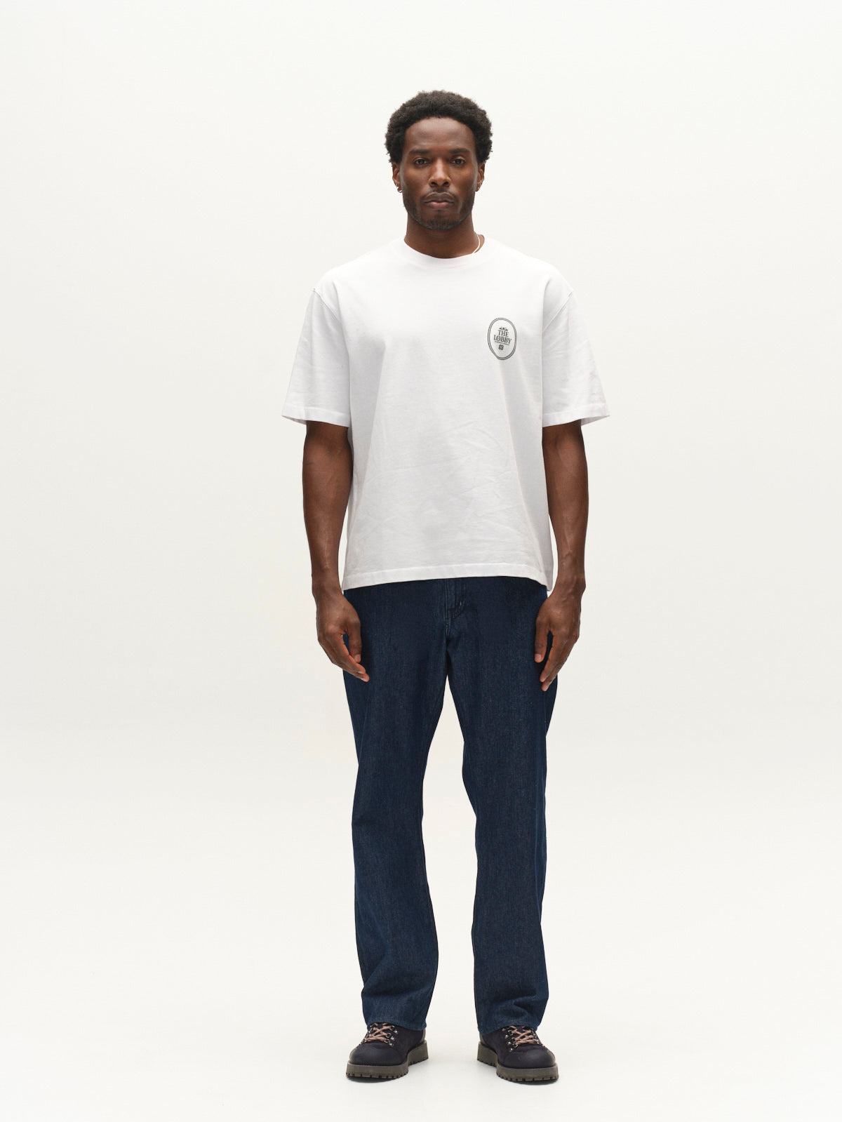 A man stands facing forward against a plain white background, wearing a white T-shirt with a small circular logo on the chest, Gabba GABnoah Tona Rinse Jeans in dark blue denim with a loose straight fit, and dark shoes.