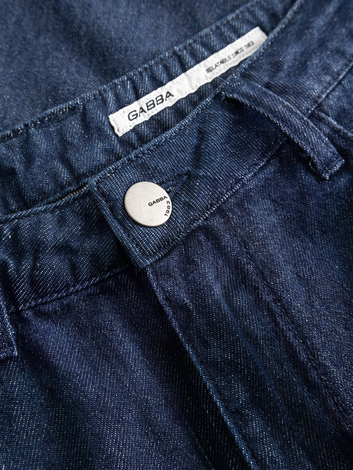 Close-up of the GABnoah Tona Rinse Jeans in dark blue denim, featuring a silver button engraved with Gabba 1983 and a white label with the brand name Gabba sewn inside the waistband. Crafted from organic cotton, these jeans showcase quality craftsmanship and sustainable materials.