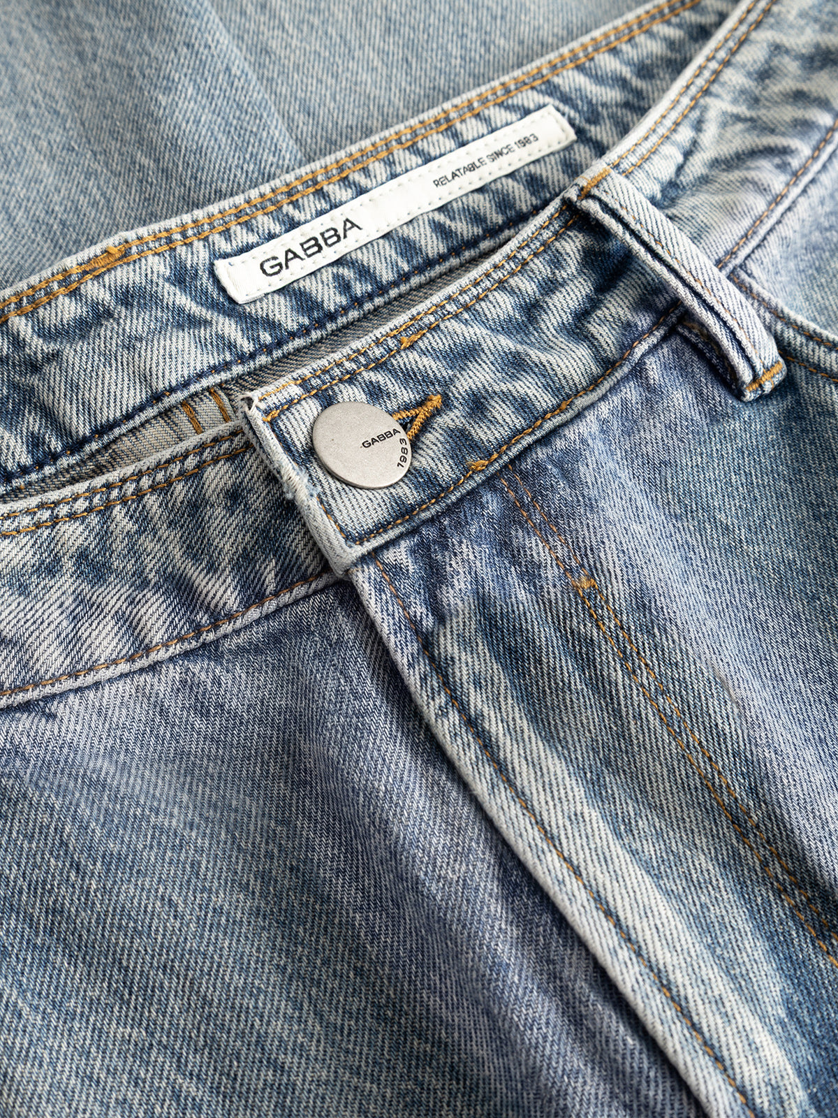 Close-up of the GABmax Tona RS2100 Jeans in Mid Blue Denim by Gabba, featuring a visible button and waistband. The label reads “Gabba.” Crafted from organic cotton denim, these jeans display classic faded patterns and detailed stitching that highlight their modern straight fit design.